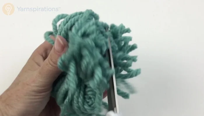 Trim the loops of the pom-pom, carefully avoiding the long tie yarn. Attach the pom-poms to the top of the hat using the long tie yarn.