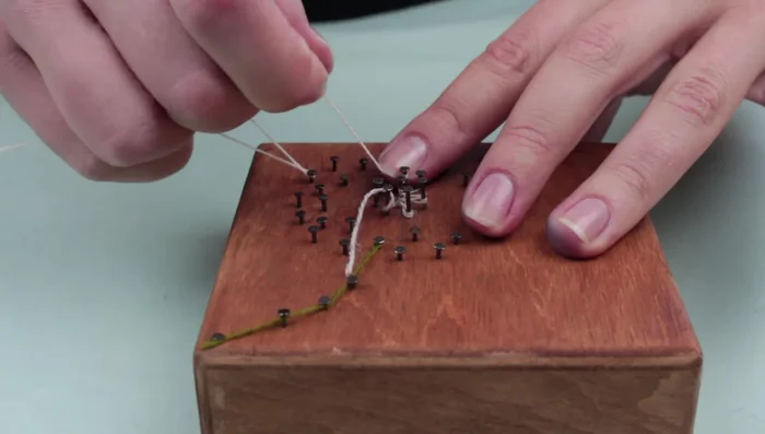 Weave white thread around the nails to create the dandelion flower, starting from the center and moving outwards. Use tweezers to help press the thread down.