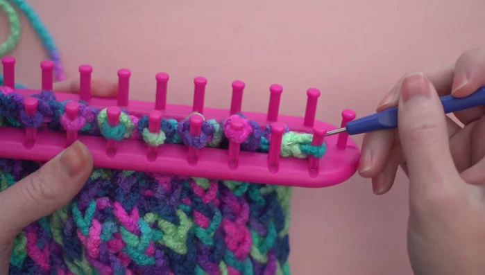 Creating a loop for the cast-off edge by moving the loop on the second peg to the first, taking the bottom loop off with the hook, and repeating until there is just one loop left.